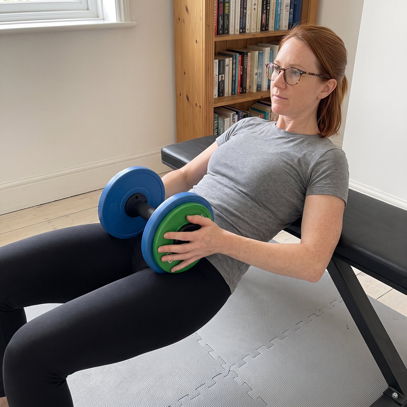 A woman in a grey t-shirt and glasses doing a hip thrust off a black bench in a spare room, holding one dumbbell handle loaded with blue and green plates across her hips