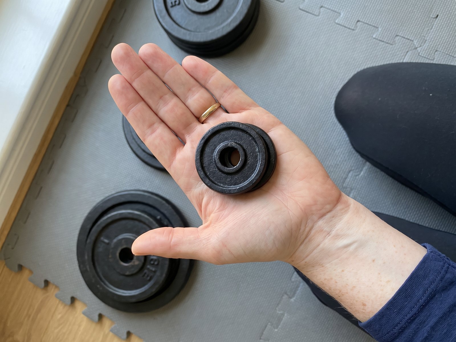 Looking down at a woman's open palm holding two small black iron weight plates above a grey foam mat, larger plates on the mat below