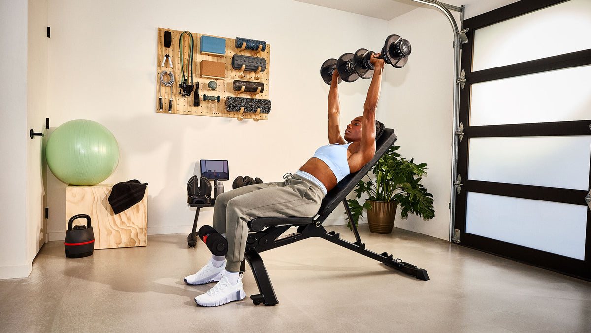 A woman on an incline bench in a garage gym pressing two black Bowflex SelectTech dial dumbbells overhead