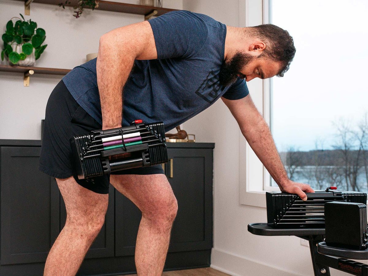 A man in a living room doing a one-arm row with a black PowerBlock Elite USA block dumbbell, its stand beside him