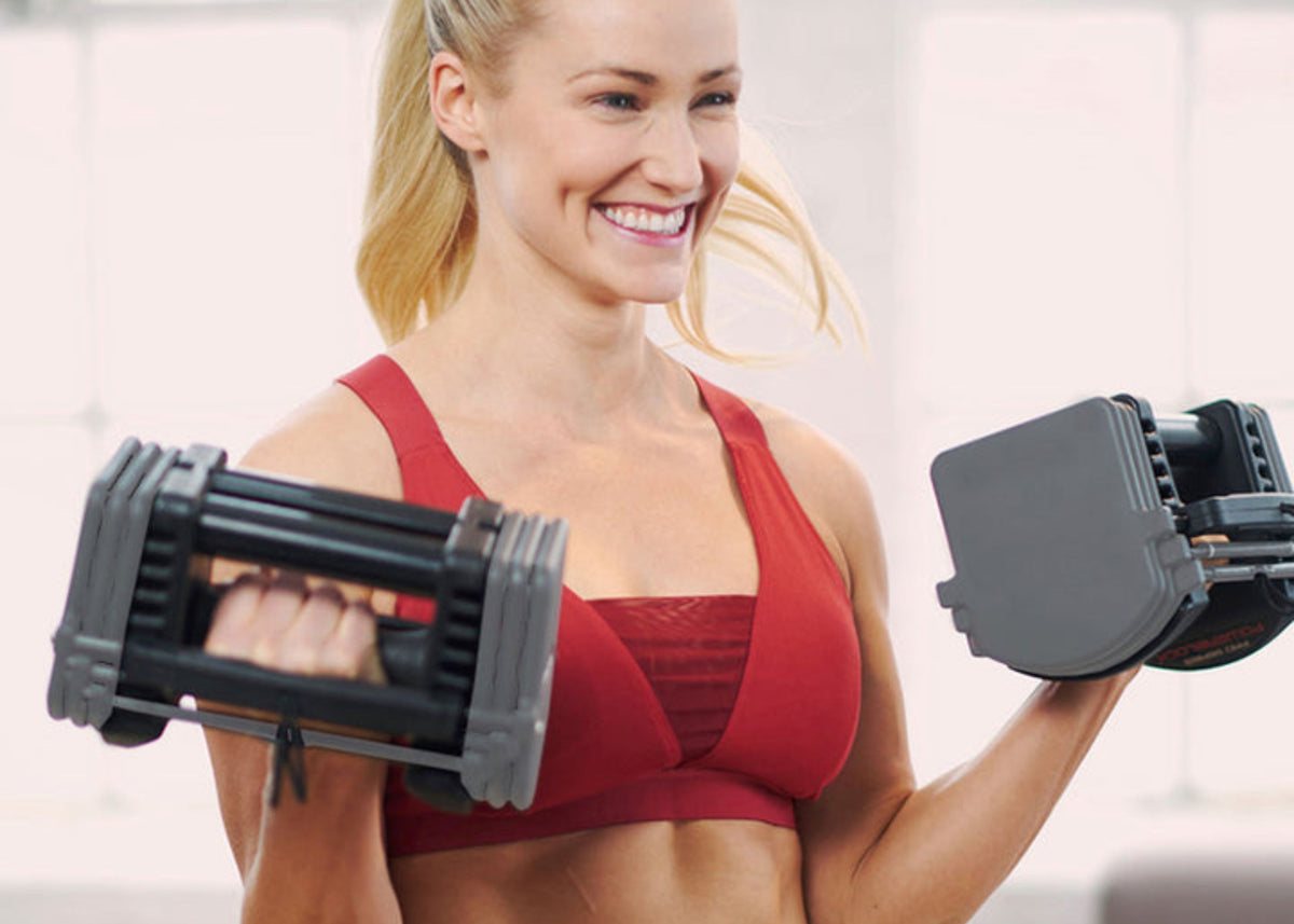 A smiling woman in a red sports top curling two grey PowerBlock Sport 24 block dumbbells
