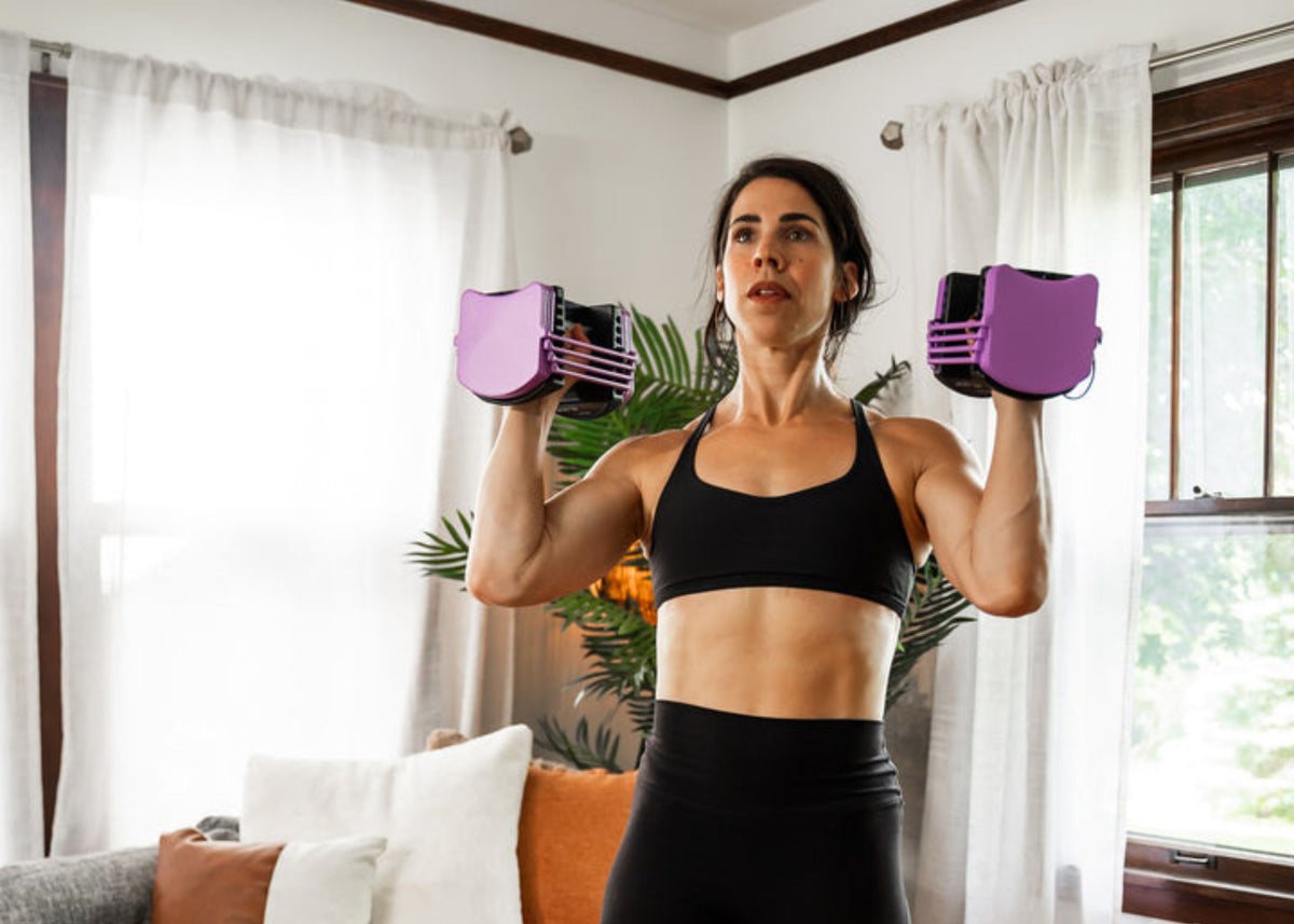 A woman in a living room pressing two lavender PowerBlock Sport 24 dumbbells overhead
