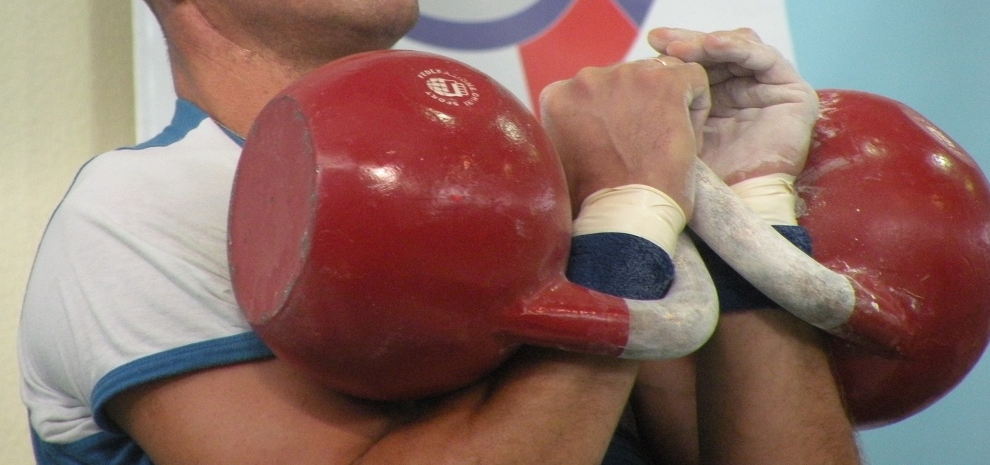Two heavy competition kettlebells held in the front rack against a lifter's forearms, hands chalked and wrists taped