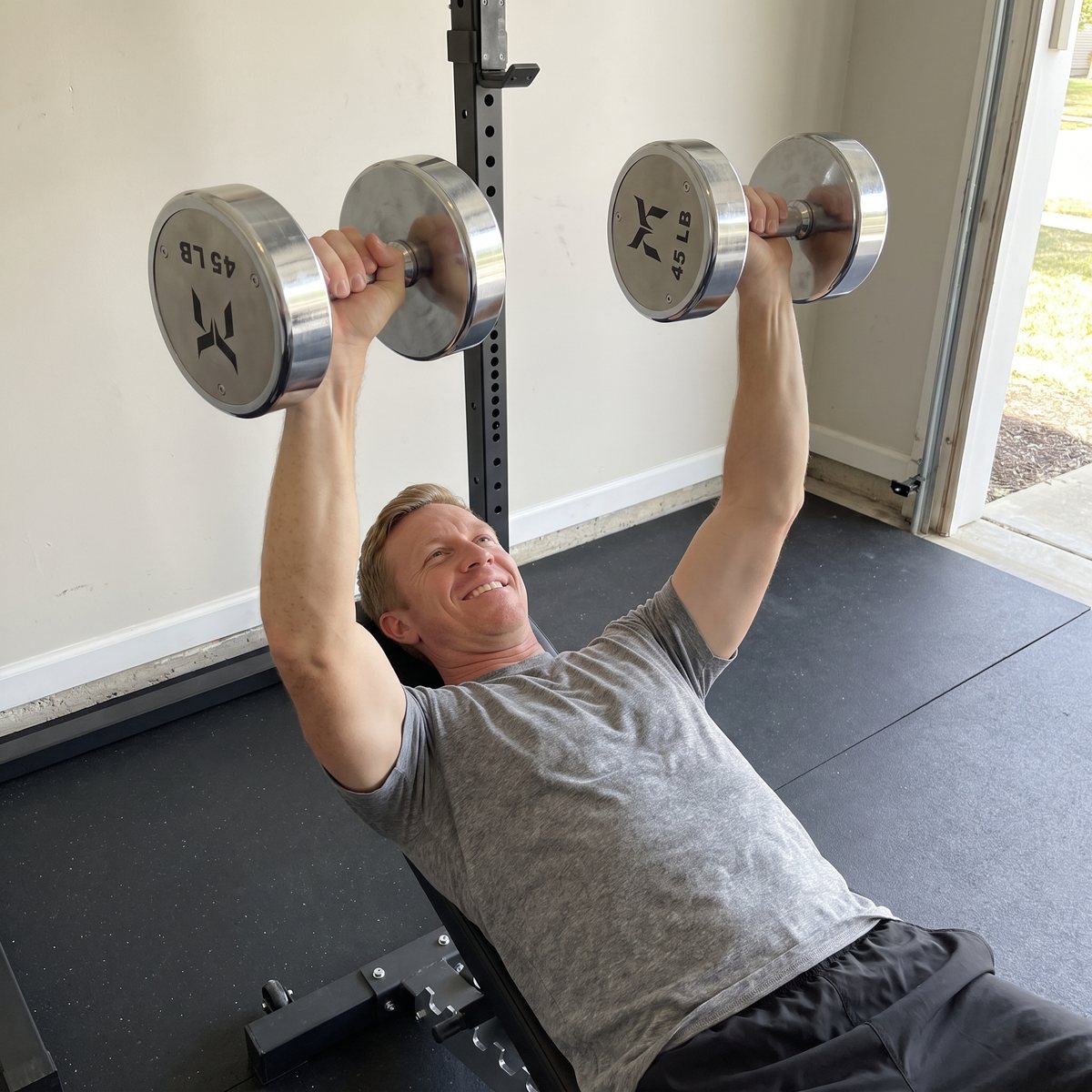 A man lying on a bench in a garage gym pressing two polished chrome dumbbells with 45 LB marked on the end caps