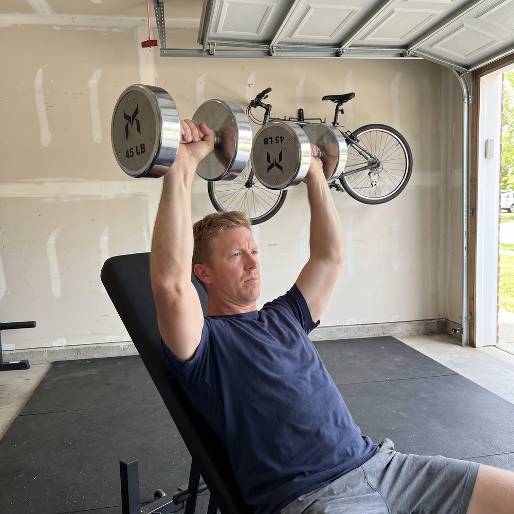 A man in a navy t-shirt pressing two chrome 45 lb dumbbells overhead on an incline bench in a garage with a bicycle hanging on the wall