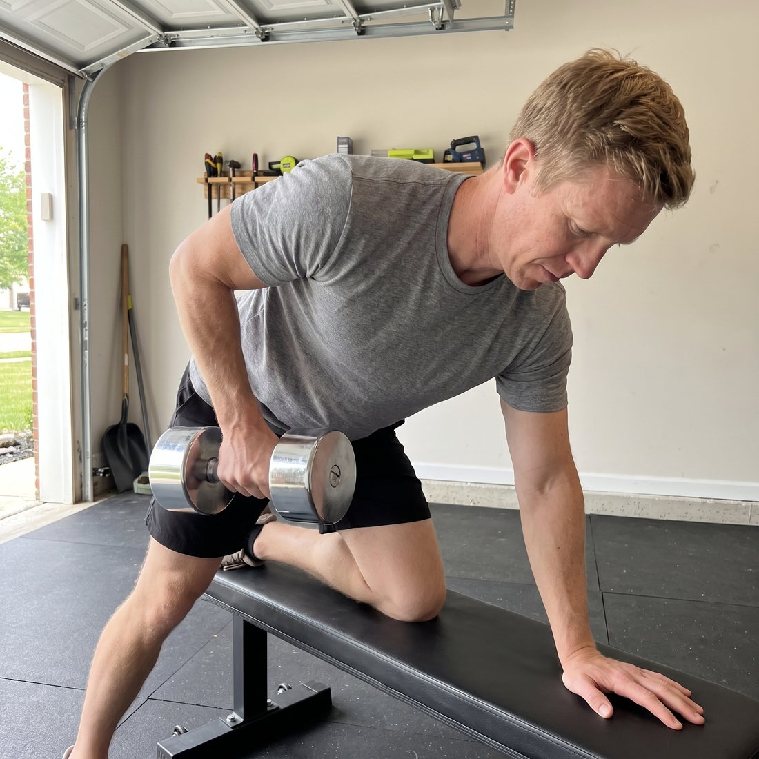 A man in a grey t-shirt doing a one-arm row with a chrome dumbbell, one knee on a flat bench in his garage gym