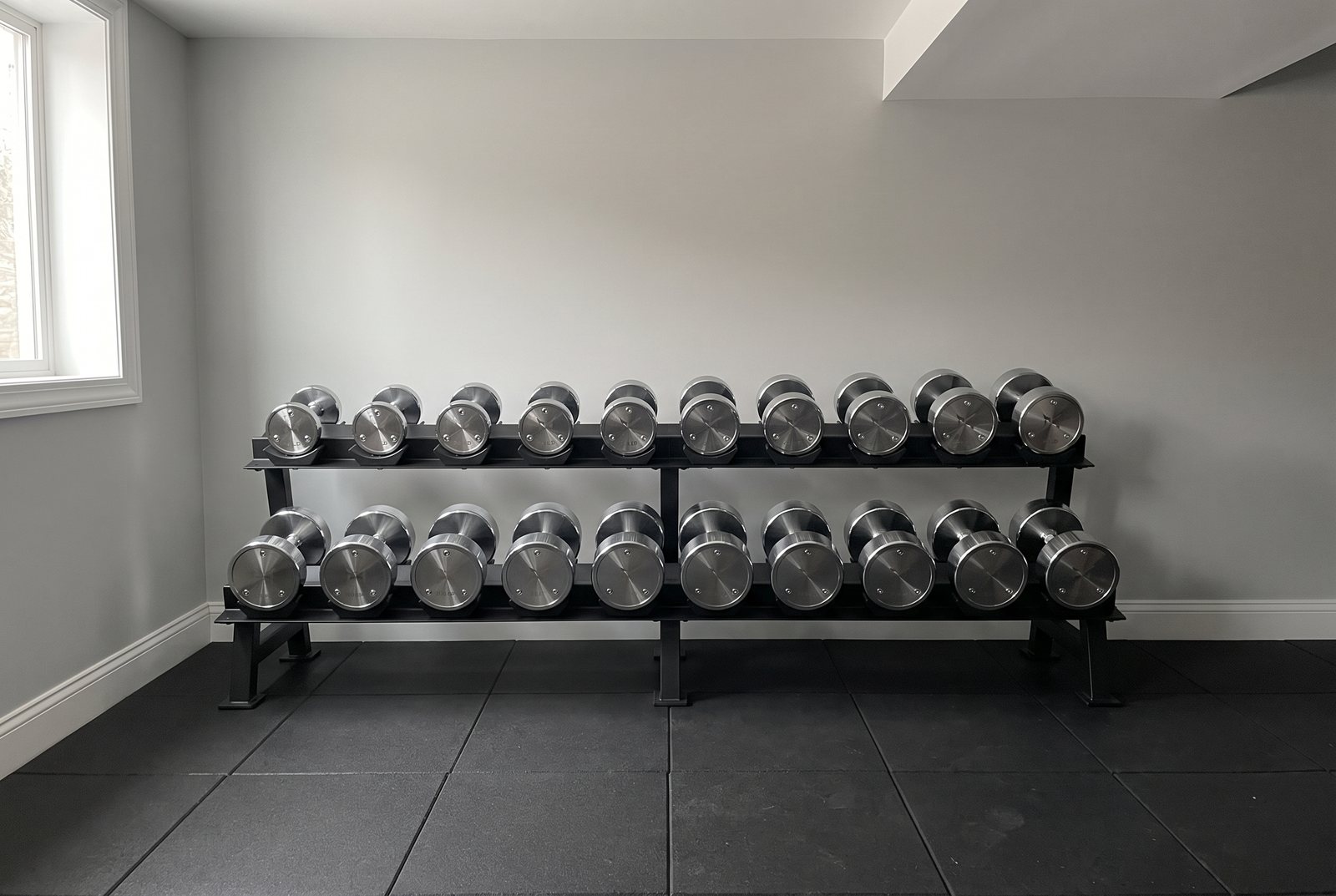Ten pairs of polished chrome dumbbells on a black two-tier rack against a light grey wall in a basement home gym