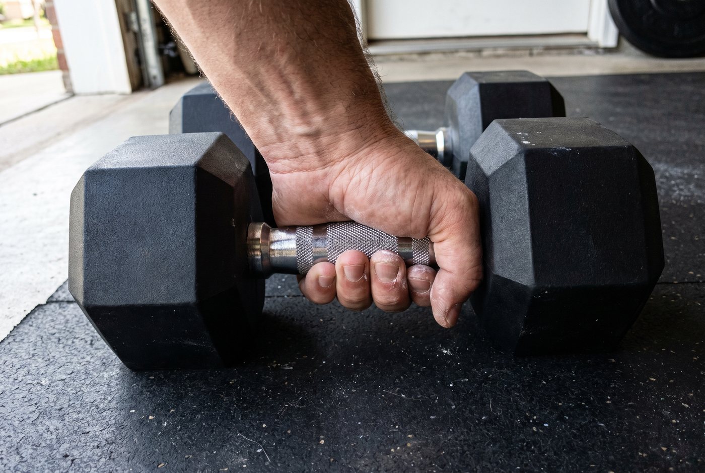 Close-up of a hand gripping the diamond-knurled steel handle of a black rubber hex dumbbell on a garage mat, a little chalk in the knurl