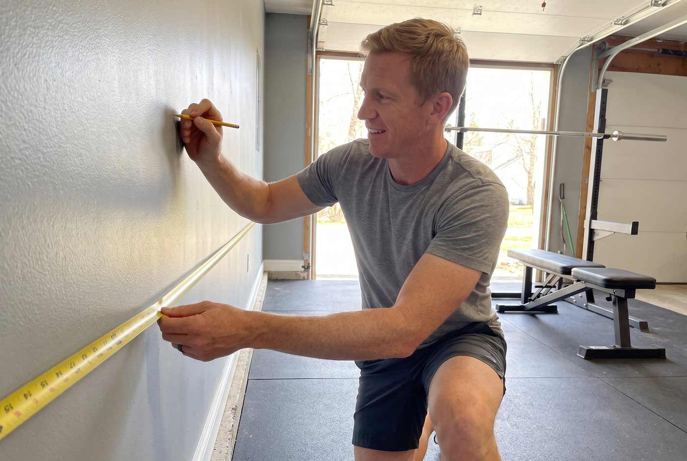 A man kneels on rubber mats in his garage and holds a yellow tape measure against a bare wall where a dumbbell rack will go