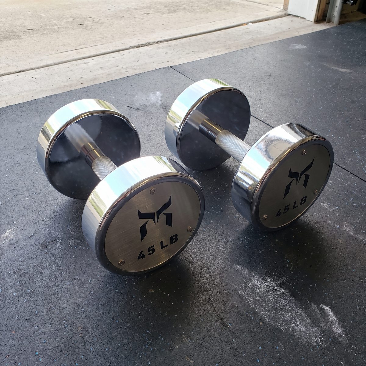 Two chrome 45 pound dumbbells on a rubber garage mat, with the weight and logo recessed into the end caps