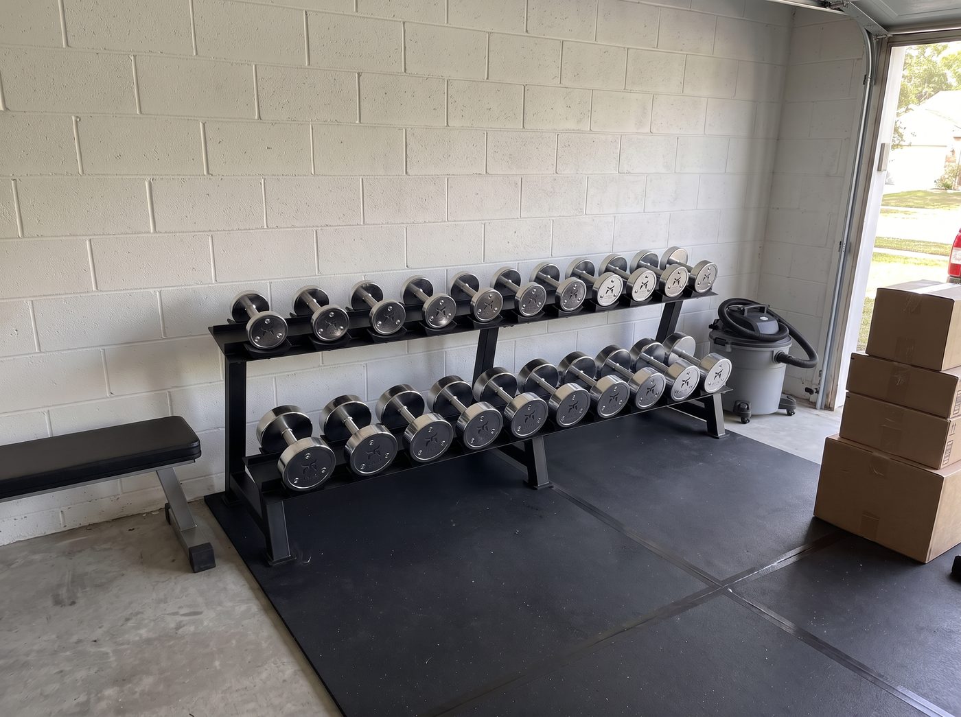 A rack of ten pairs of chrome dumbbells on two shelves against a garage wall, on black rubber mats