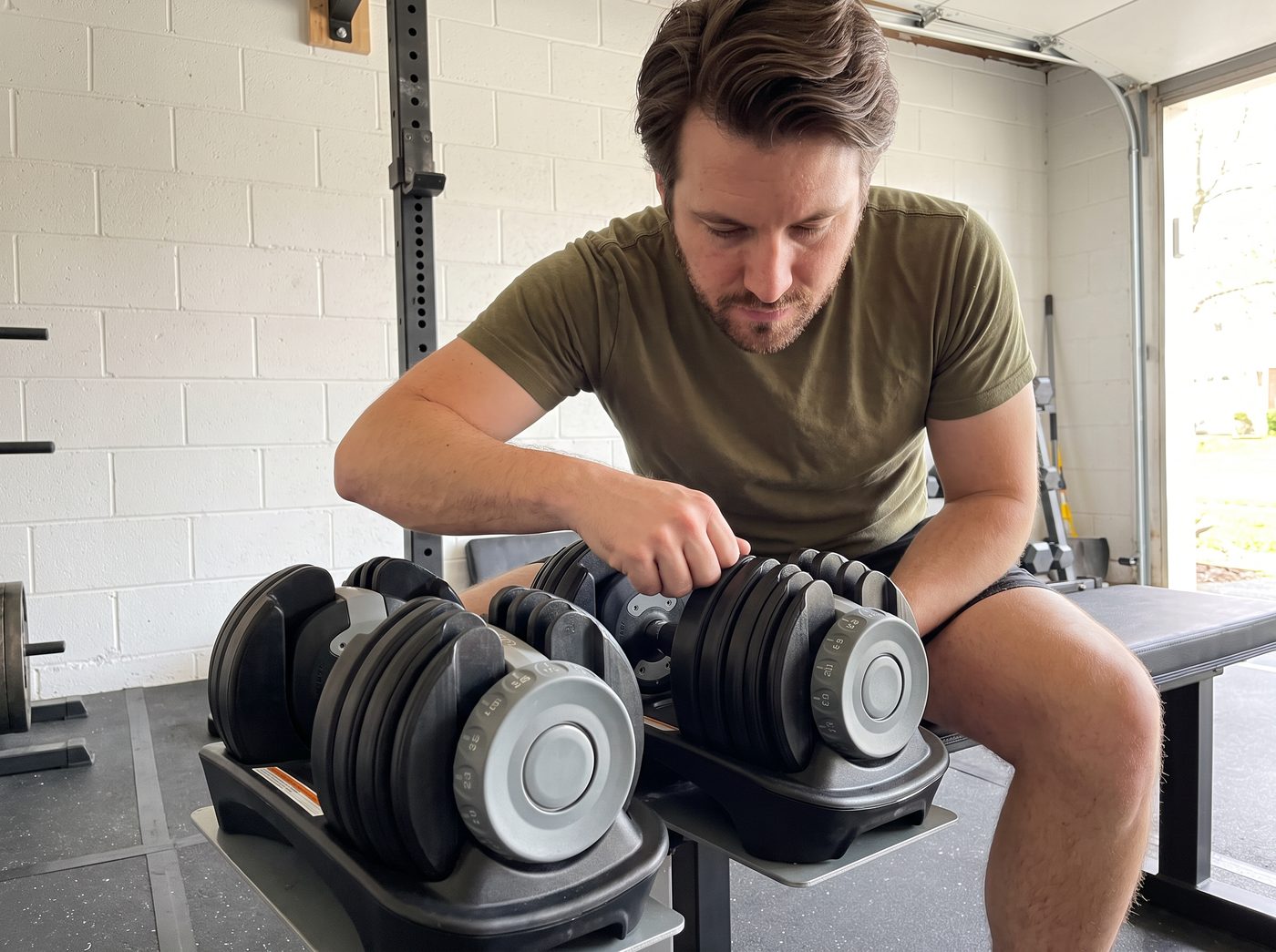 A man on a bench turning the dial of an adjustable dumbbell resting in its plastic cradle