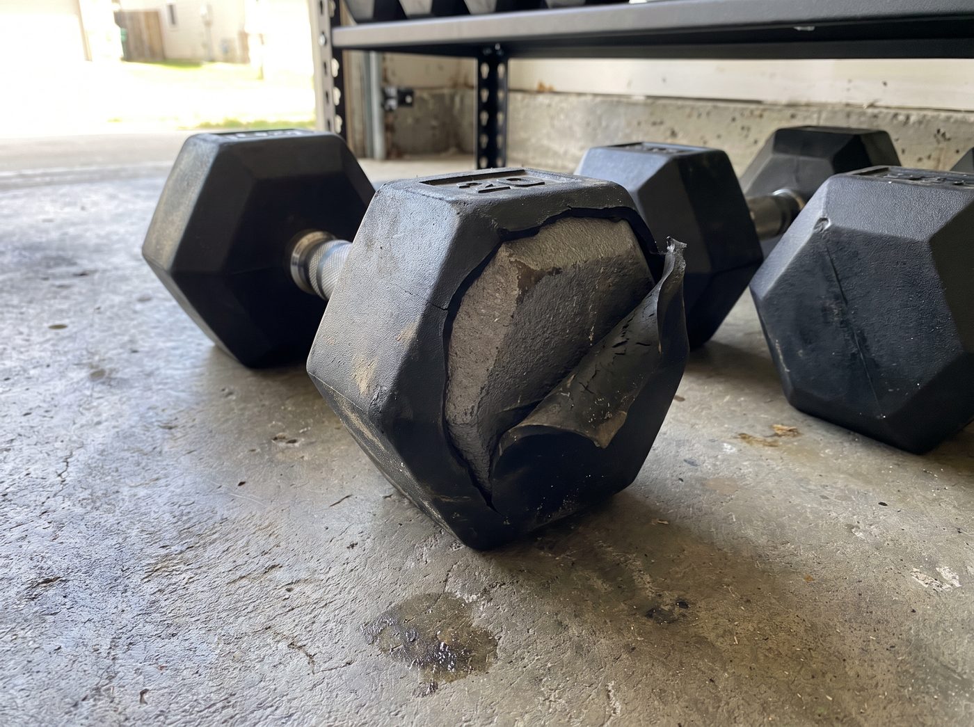 A rubber hex dumbbell on a concrete garage floor with the rubber torn open at one corner, showing the iron underneath