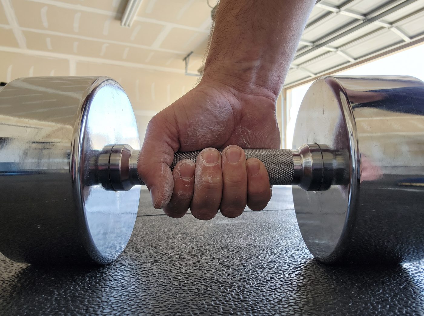 A chalked hand gripping the knurled handle of a chrome dumbbell resting on a rubber mat