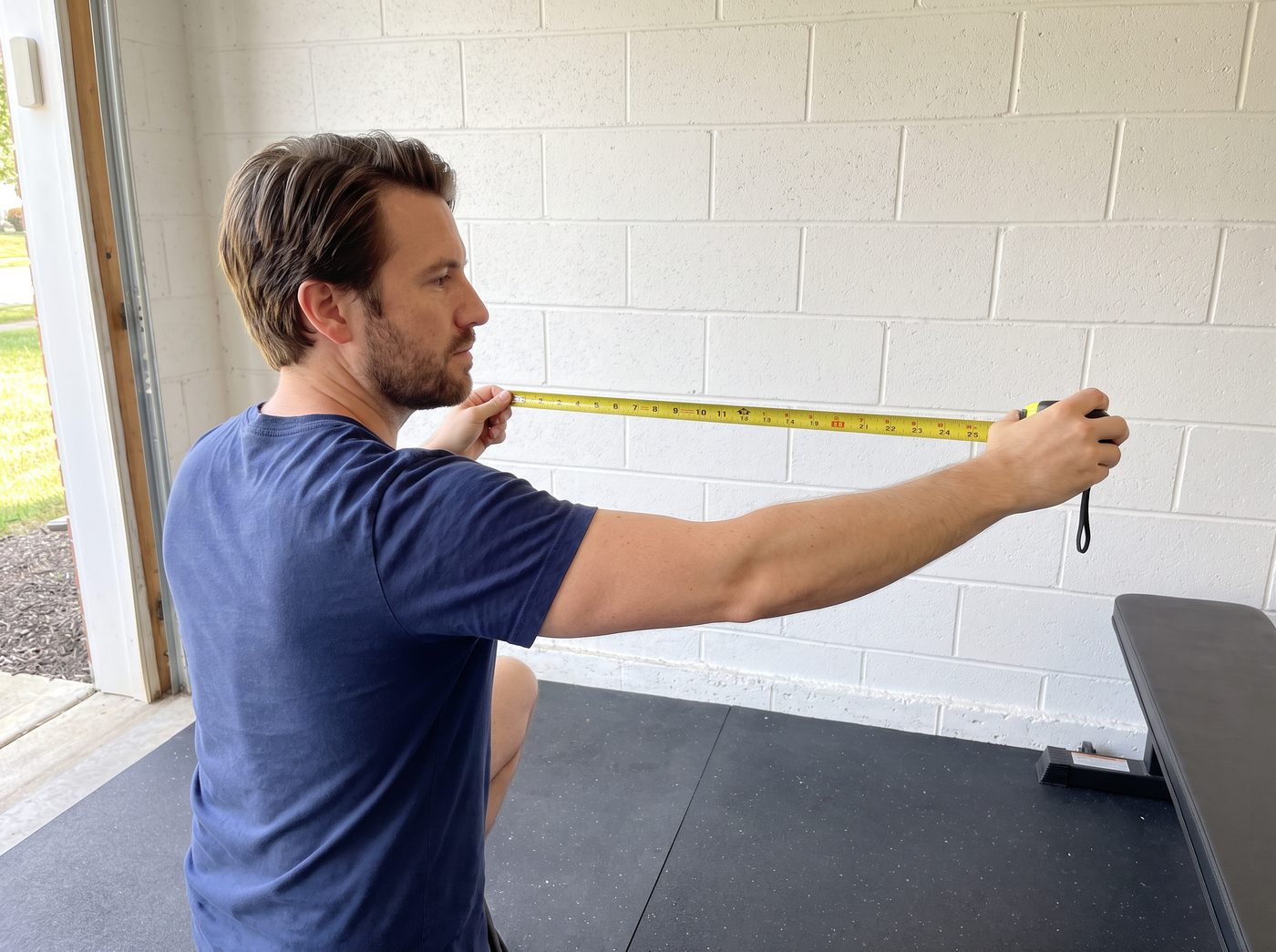A man stretching a yellow tape measure along an empty garage wall above rubber floor mats