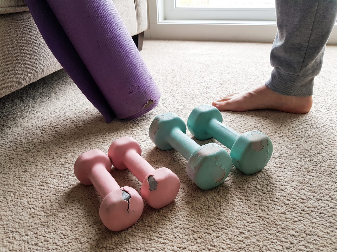 Two pairs of worn pink and teal neoprene dumbbells on a living room rug next to a rolled purple yoga mat
