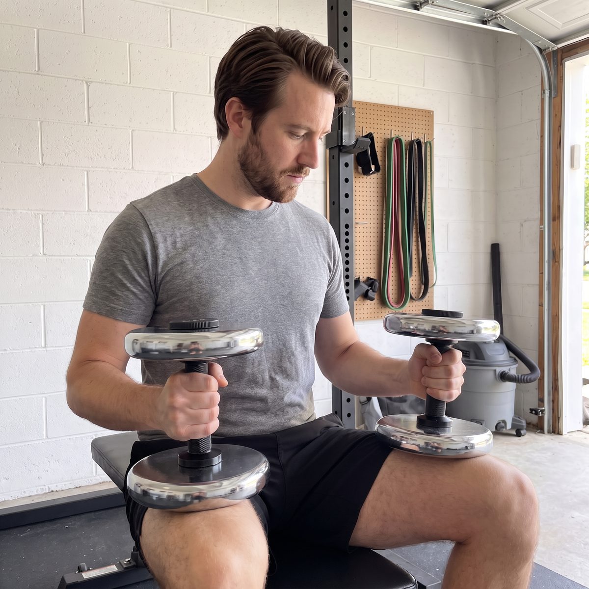 A man seated on a bench in a garage holding two short dumbbell handles loaded with chrome weight plates