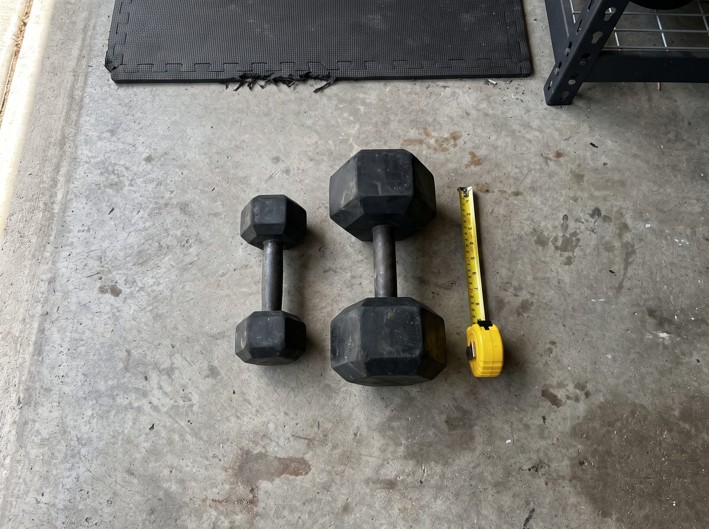 A 5 pound and a 50 pound rubber hex dumbbell side by side on a garage floor next to a yellow tape measure