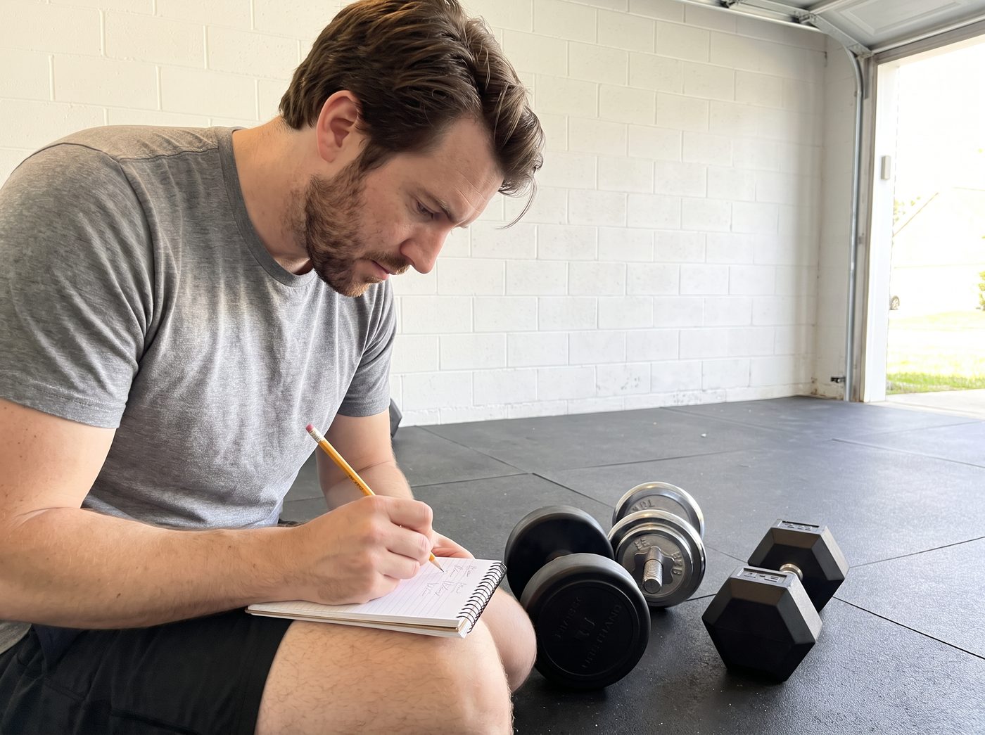 A man kneeling on a garage mat writing in a notebook beside a urethane, a chrome and a rubber hex dumbbell