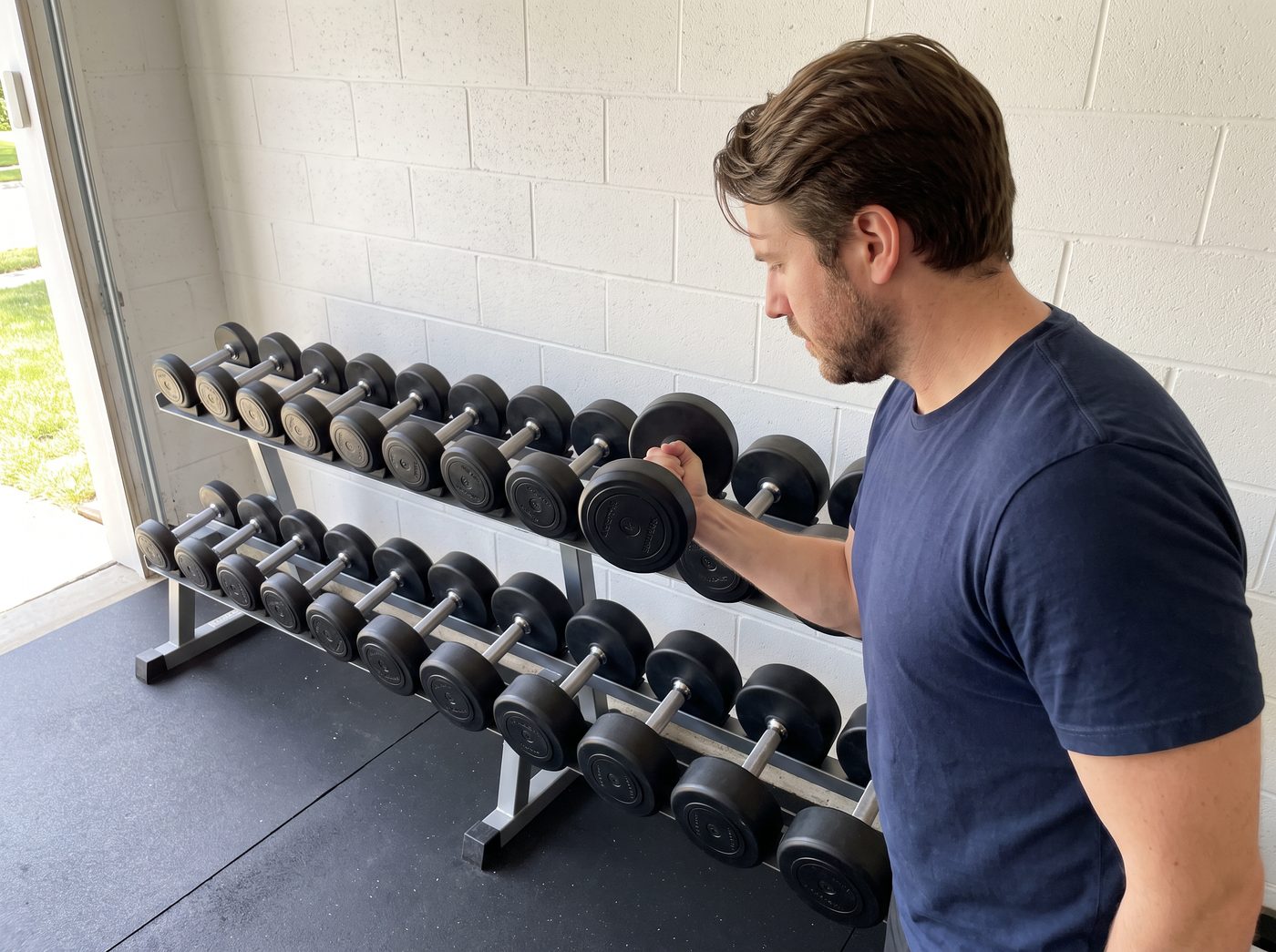 A man lifting a black round urethane dumbbell off a rack of matching urethane dumbbells in a garage