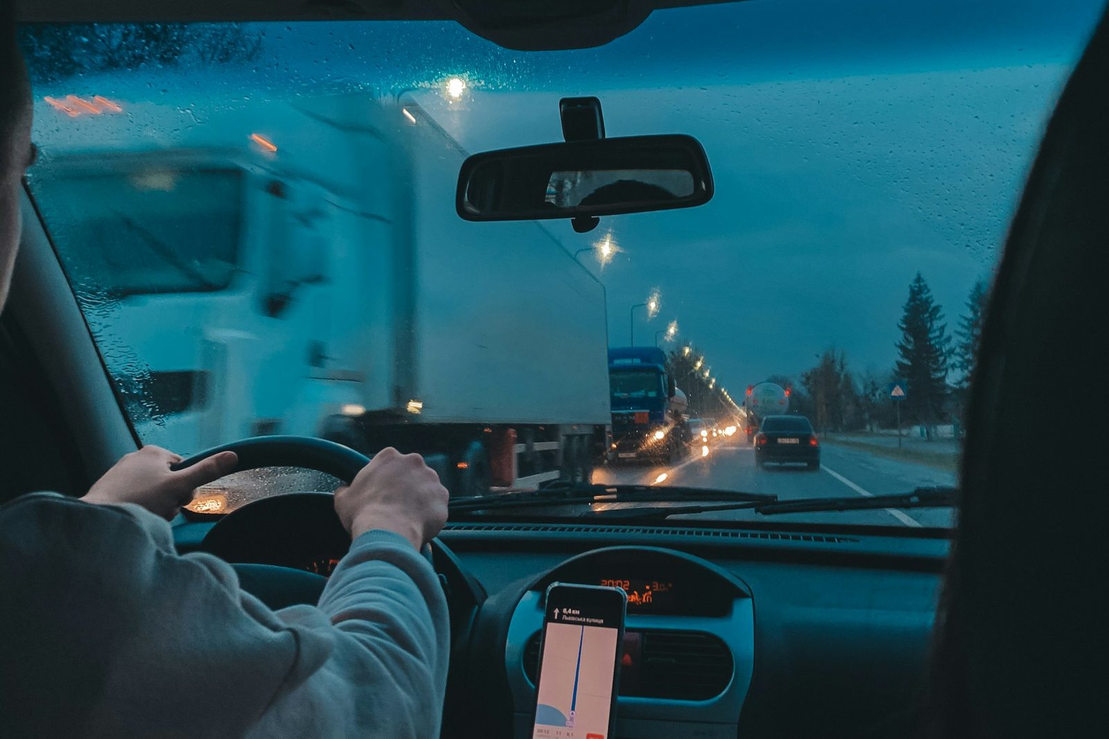 Driver's view from inside a car at dusk in the rain, hands on the wheel, a truck ahead and a phone on the dash showing a navigation route
