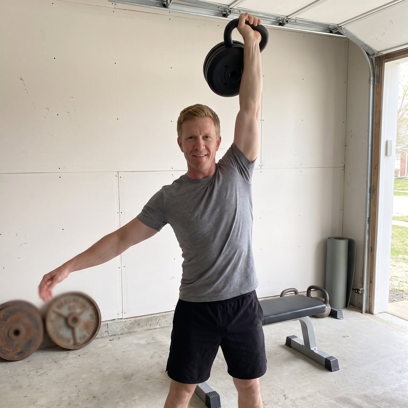 A man in a garage holding a plate-loaded kettlebell overhead with one straight arm