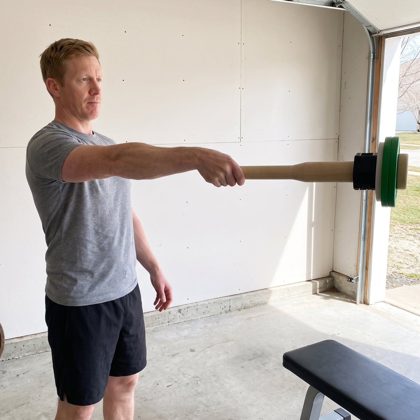 A man in a garage holding a wooden club straight out in front of him with plates loaded at the far end