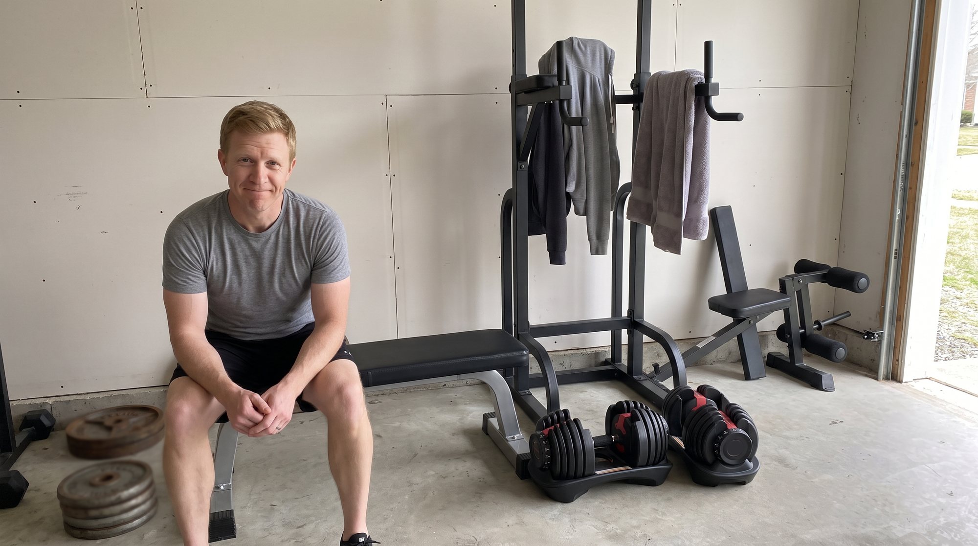 A man sitting on a weight bench in his garage with a rueful smile, next to a power tower with hoodies and a towel hanging on it, dial dumbbells in their trays and an unused leg attachment by the wall