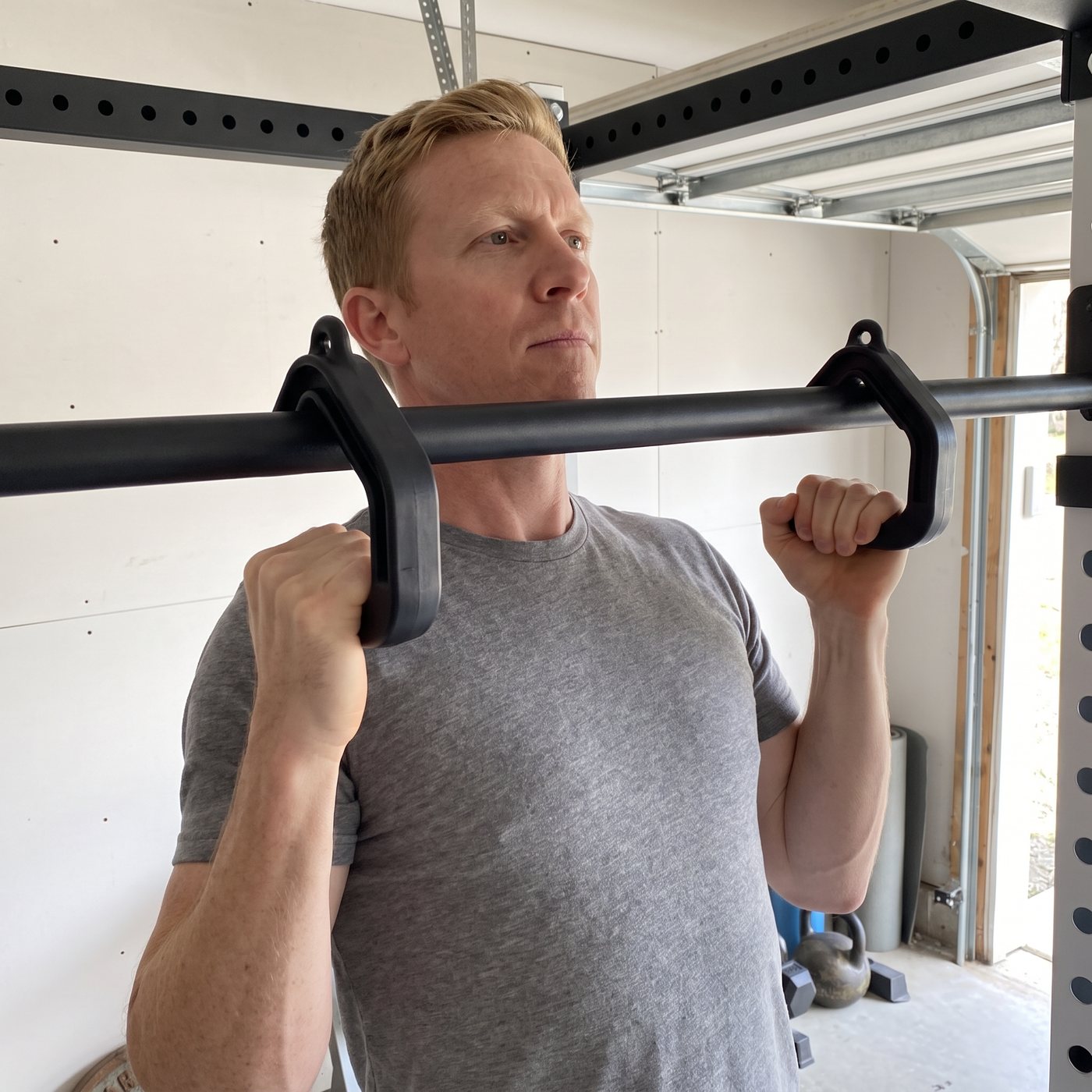 A man gripping two hook-shaped neutral-grip handles hung over the straight pull-up bar of his rack