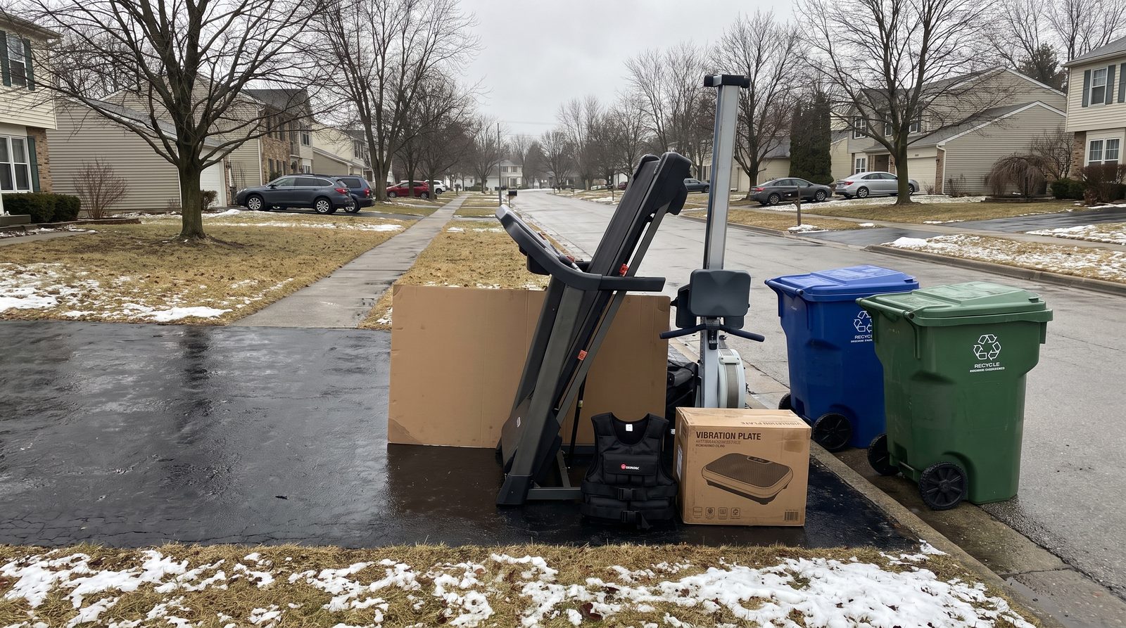 A folded treadmill, a rowing machine, a sand-filled weight vest and a boxed vibration plate left at the curb beside two recycling bins on a winter morning