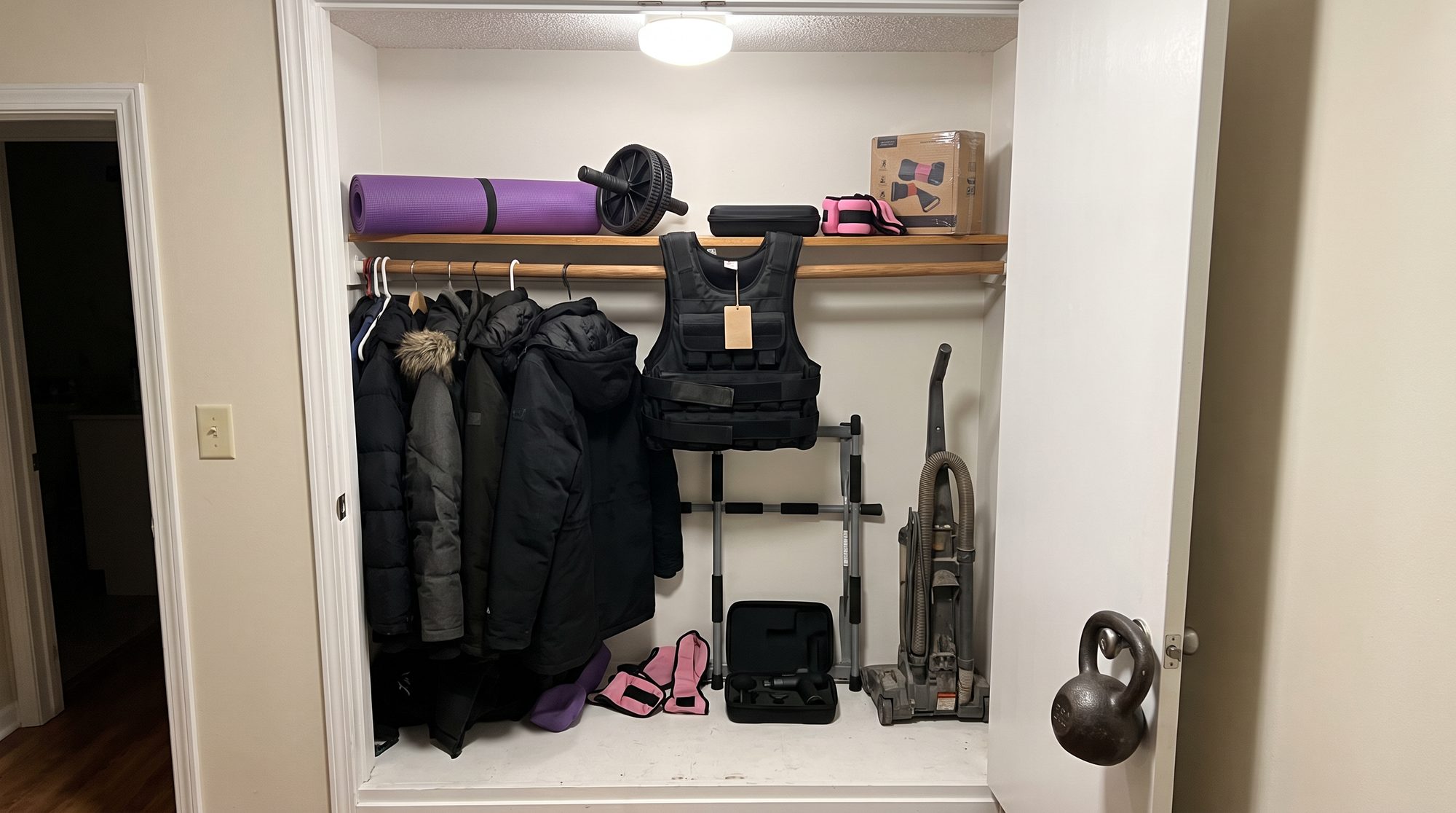 A hallway coat closet with a sand-filled weight vest, a rolled yoga mat, an ab wheel, pink ankle weights, a boxed set of resistance bands, a doorway pull-up bar and a massage gun pushed in beside the winter coats