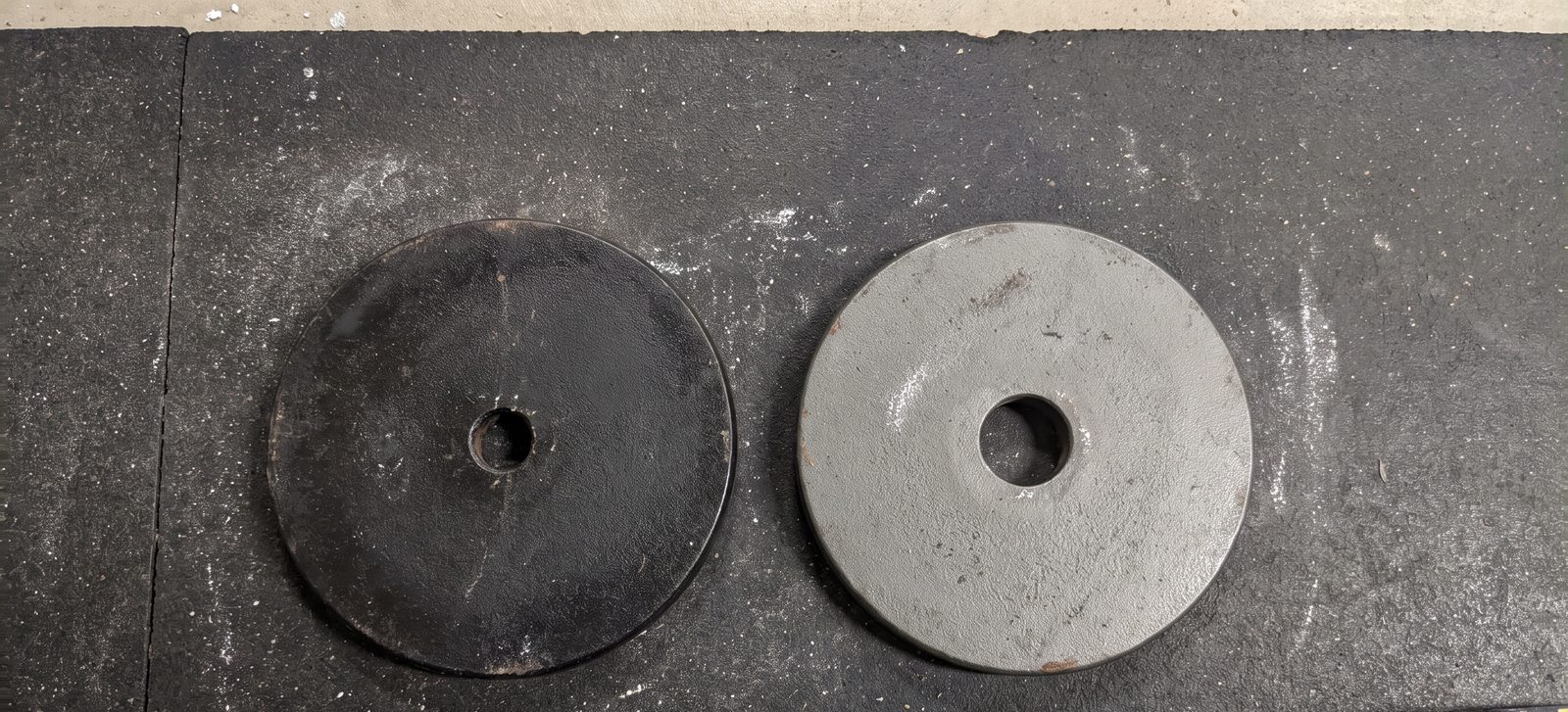 Two cast iron weight plates side by side on a rubber mat, one with a small 1 inch centre hole and one with a large 2 inch centre hole