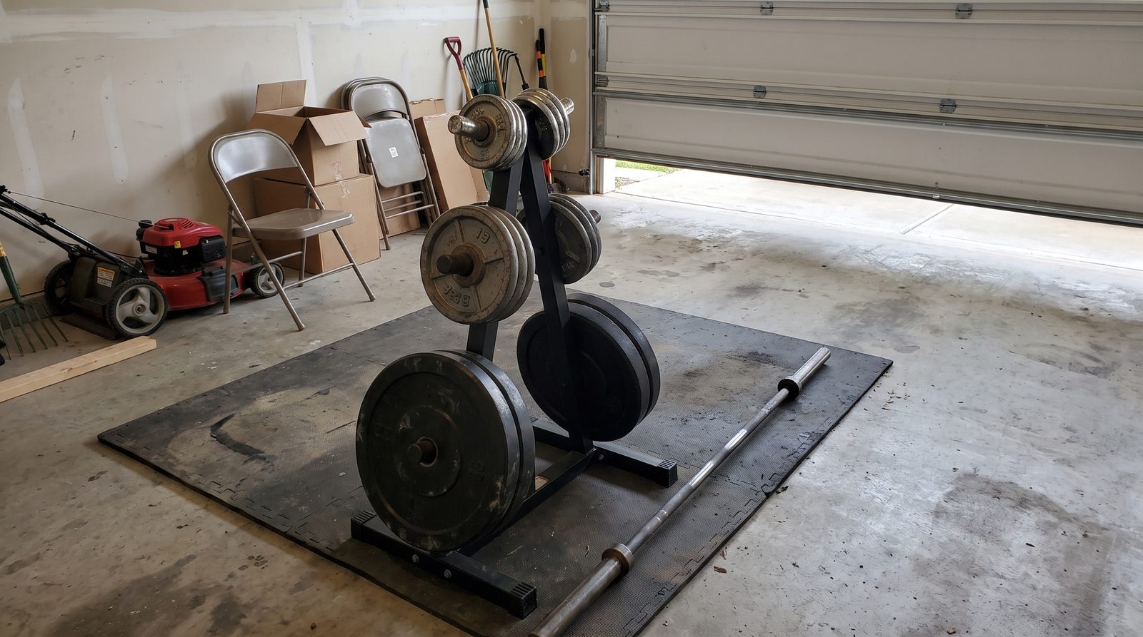 A plate tree of mismatched Olympic weight plates and a barbell on a rubber mat in the corner of a two-car garage
