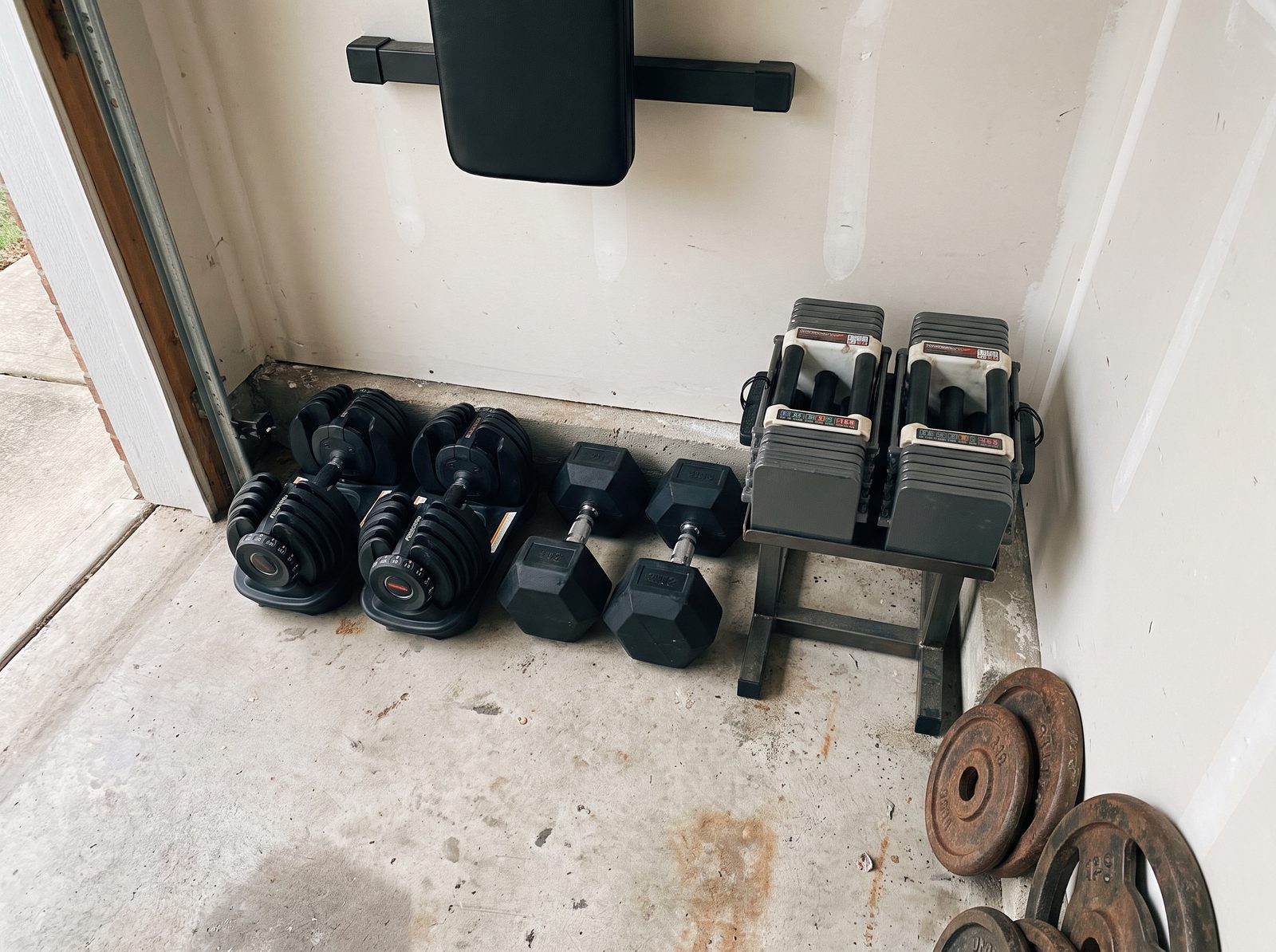 The corner of a garage gym floor with a pair of dial-adjustable dumbbells, a pair of heavy rubber hex dumbbells and a pair of quick-adjust block dumbbells on a stand, lined up against the wall