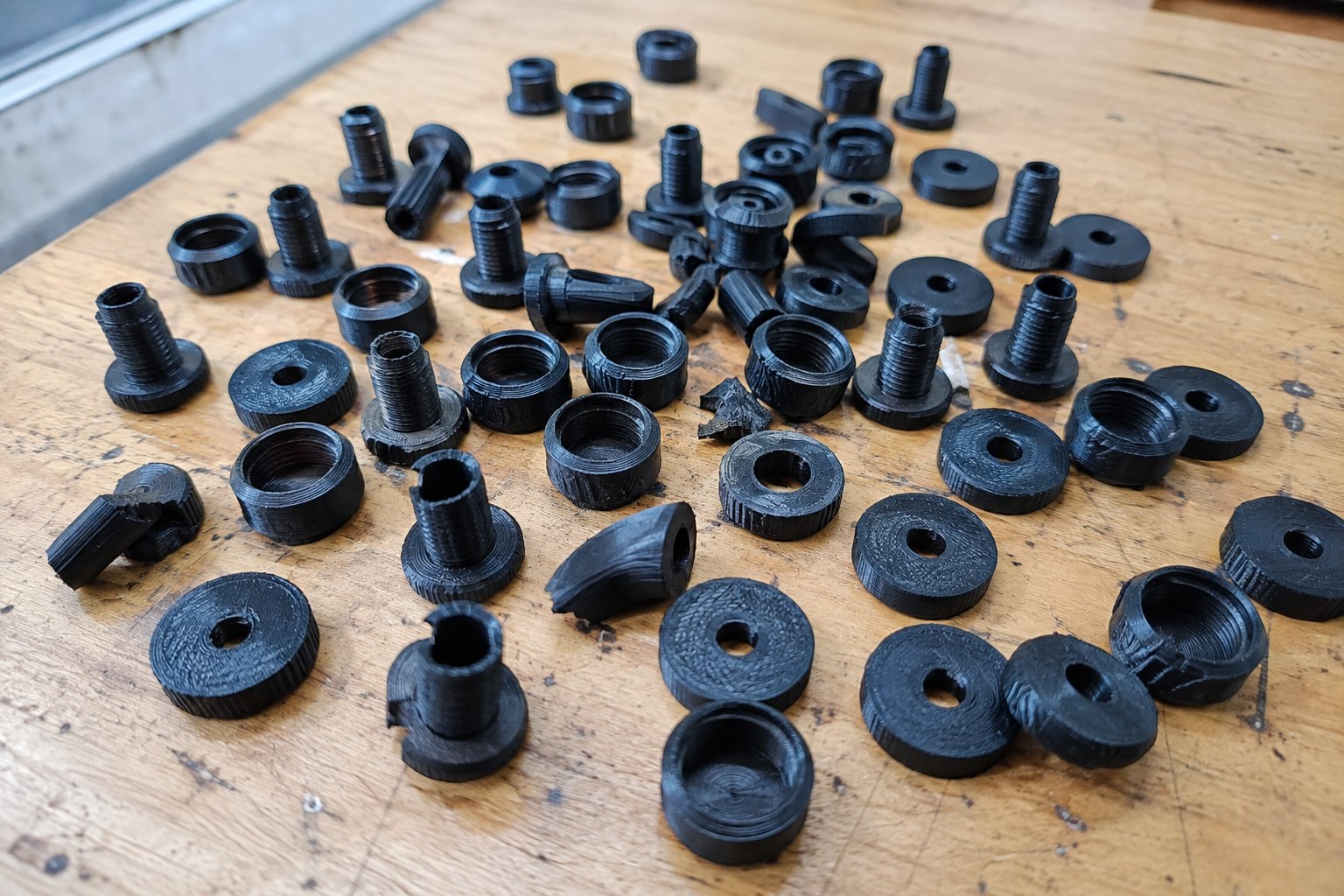 Several dozen small black 3D-printed parts spread across a workbench, threaded posts and knurled collars and flat discs, printing layer lines visible and a few of them warped or snapped