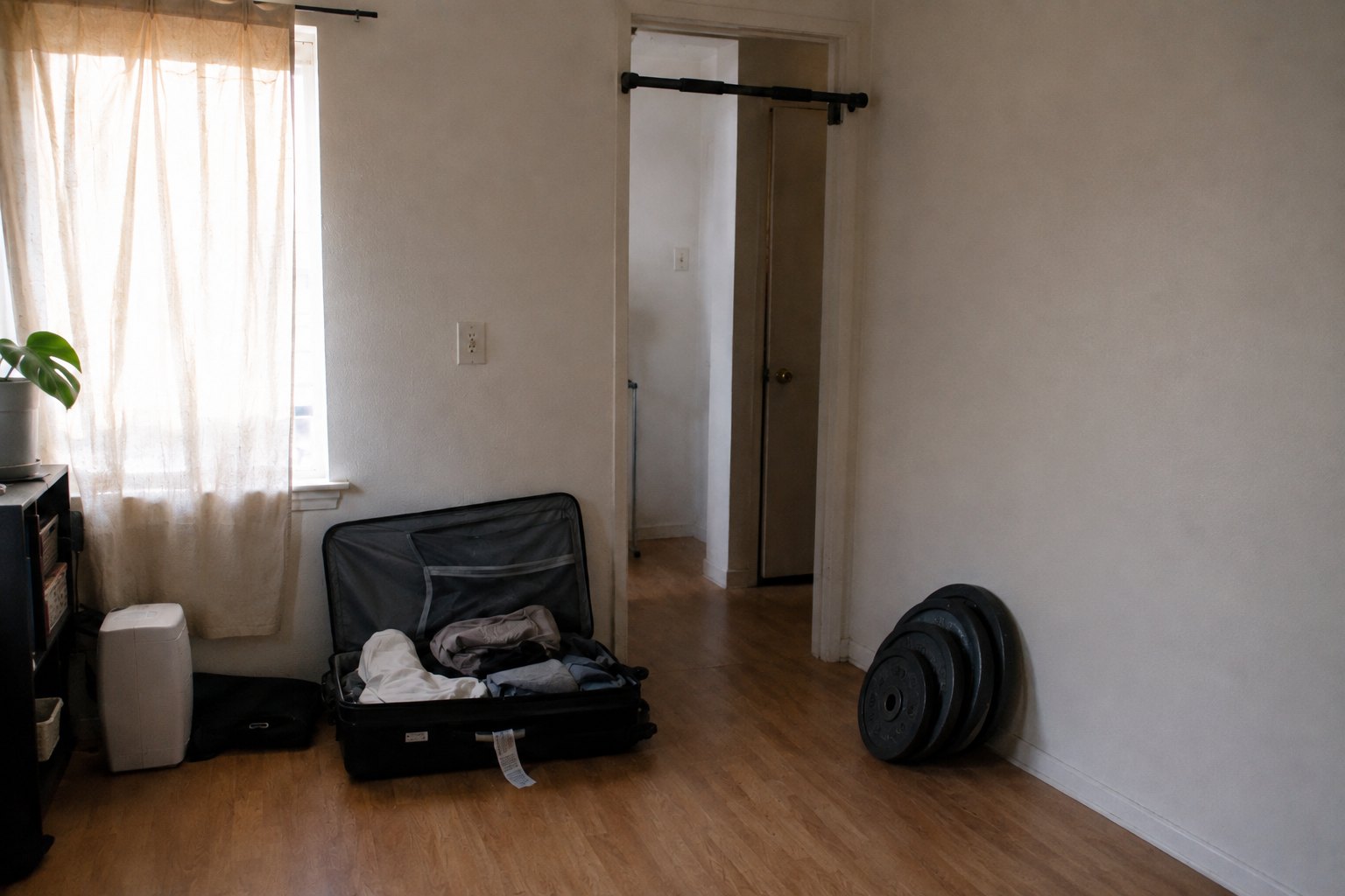 A sparsely furnished rented room used as a makeshift gym, a half-packed suitcase open on the floor, a chin-up bar wedged in the doorway and a stack of cast-iron plates leaning against the bare wall