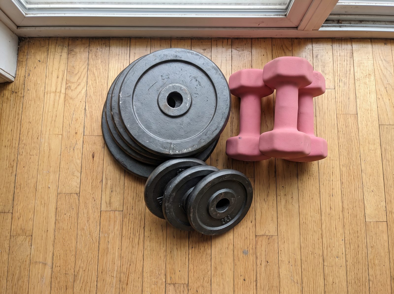 A stack of black cast iron standard weight plates on a wooden apartment floor next to two pairs of small pink neoprene dumbbells