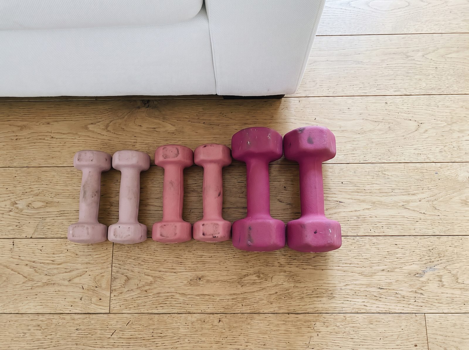 Three pairs of scuffed pink neoprene dumbbells lined up from smallest to largest on a wooden floor, with an empty space beside the largest pair