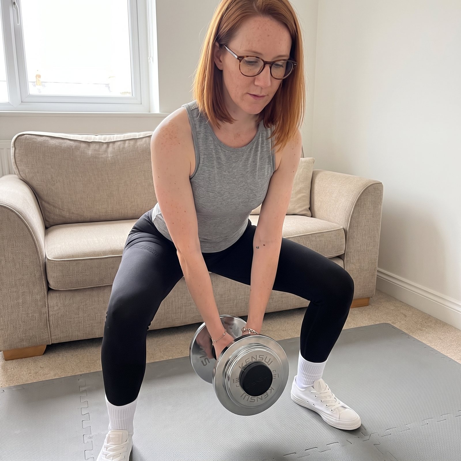 A woman in a wide stance in her living room, deadlifting a single AdaptaBELL handle held in both hands and loaded with chrome plates