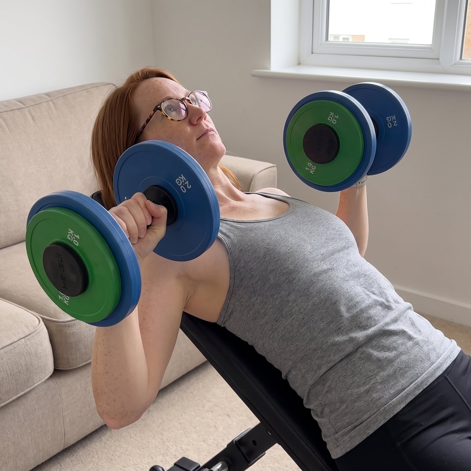 A woman doing an incline dumbbell press at home with two AdaptaBELL handles loaded with small green and blue 1 and 2 kilogram plates