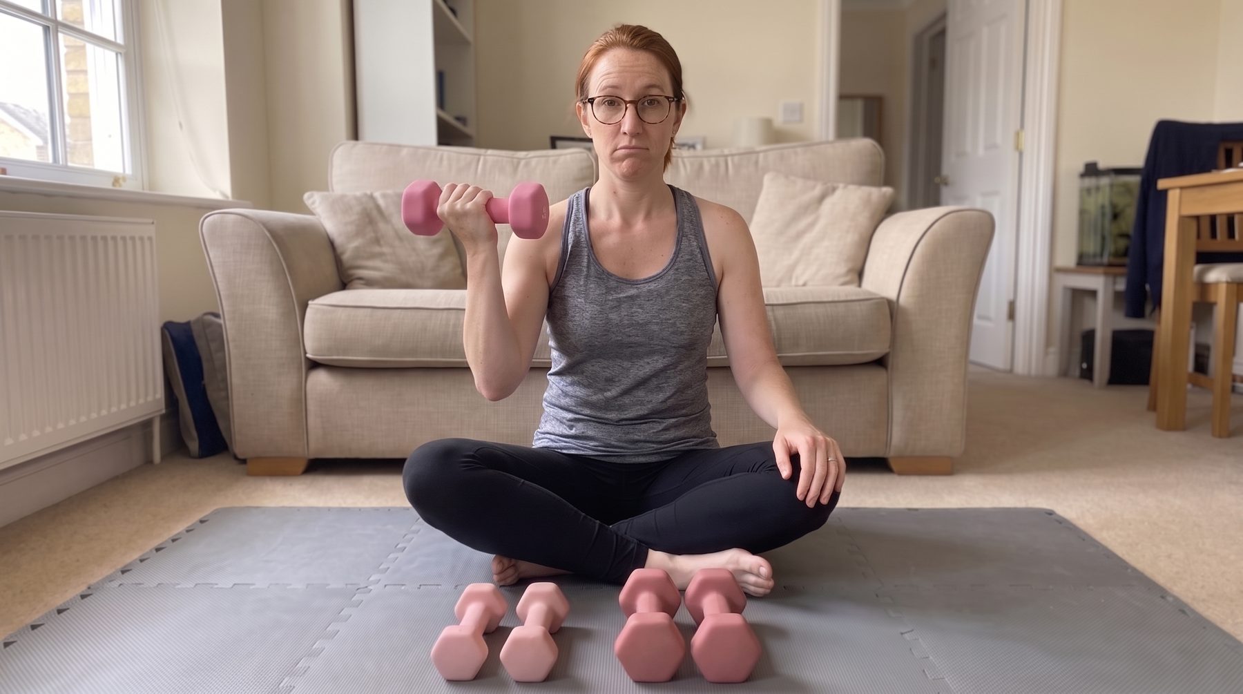 A woman sitting cross-legged on a grey mat in her living room, holding up a pink neoprene dumbbell with an unimpressed look, two smaller pairs of pink dumbbells lined up in front of her
