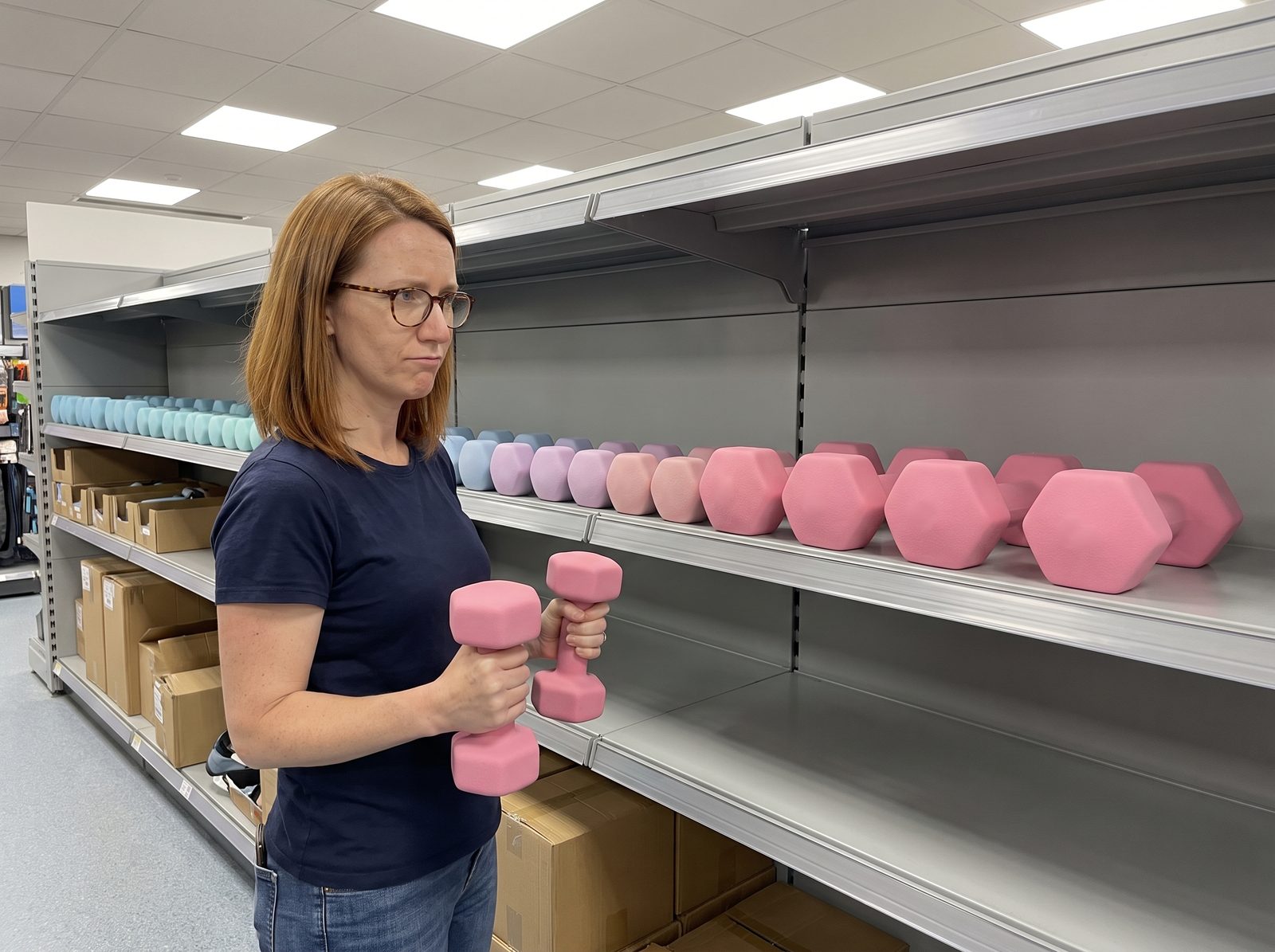 A woman in a store aisle holding a pair of pink neoprene dumbbells, looking at a shelf of pastel neoprene dumbbells that ends at her pair with bare shelf beyond it