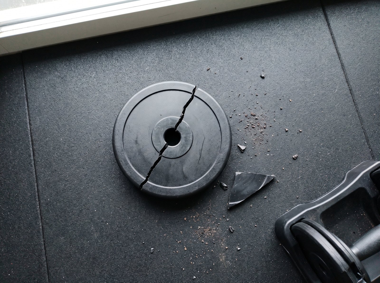 A moulded plastic weight plate from an adjustable dumbbell lying cracked in two on a rubber gym mat with a broken piece and plastic crumbs beside it