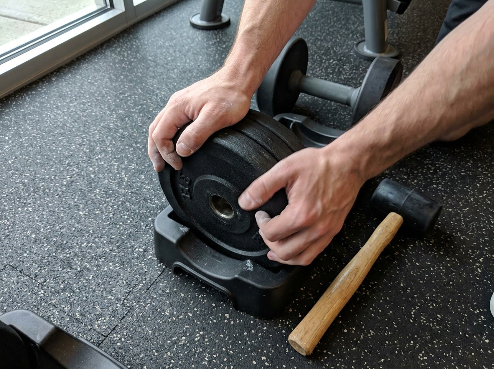 Two hands pressing a stack of black weight plates down into the slot of a plastic adjustable dumbbell cradle, with the plates sitting visibly crooked and a rubber mallet on the mat alongside