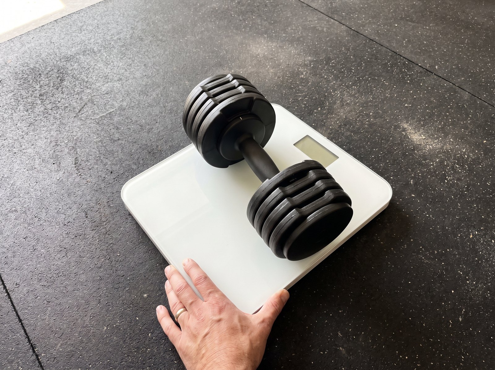 An adjustable dumbbell sitting on a flat bathroom scale on a gym floor with a hand steadying the scale