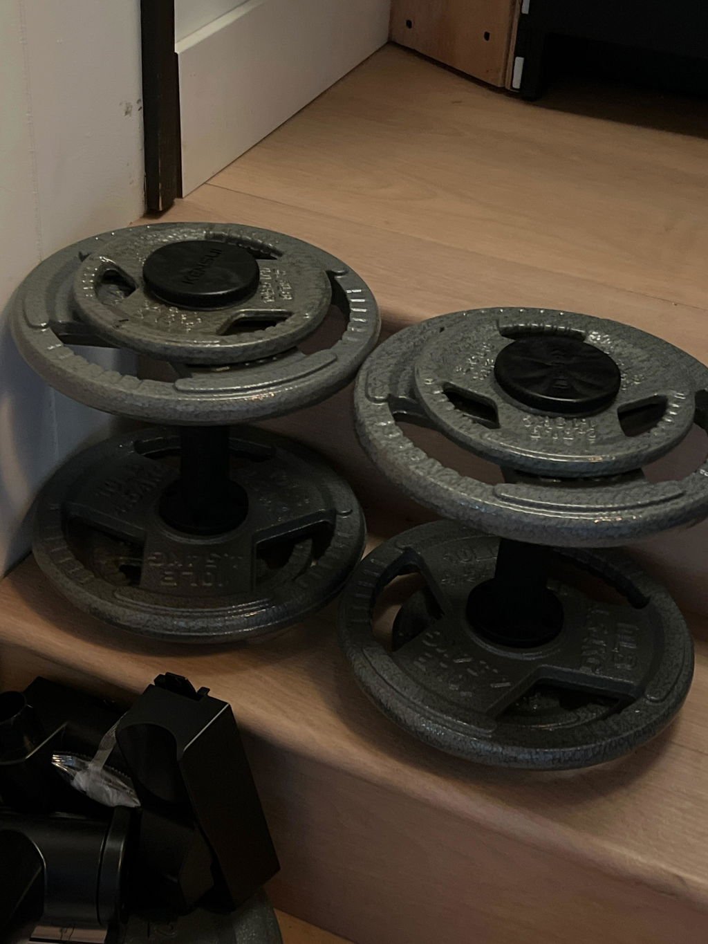 An owner's two black dumbbell handles loaded with large grey iron plates on a wooden floor at home