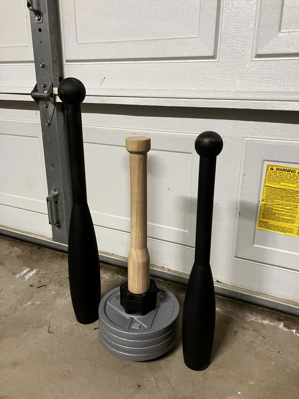 An owner's wooden plate-loaded club loaded with four grey plates, standing between two fixed black steel clubs against a garage door