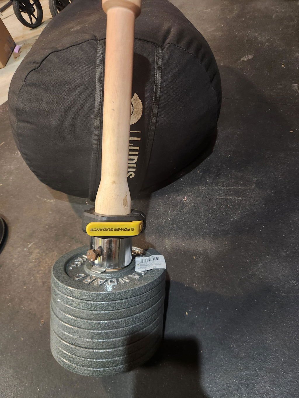 An owner's wooden plate-loaded club standing on a tall stack of grey plates next to a heavy bag in a home gym