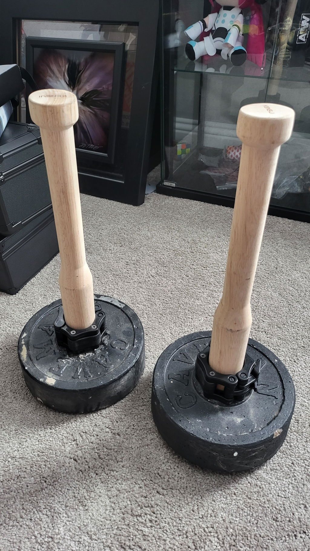 Two wooden plate-loaded clubs standing upright on black plates on a carpeted floor at home