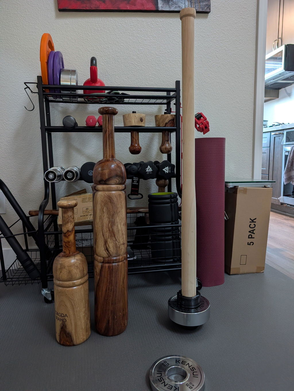 A wooden EZ-MACE loaded with a chrome plate standing beside a set of Persian clubs in a home gym